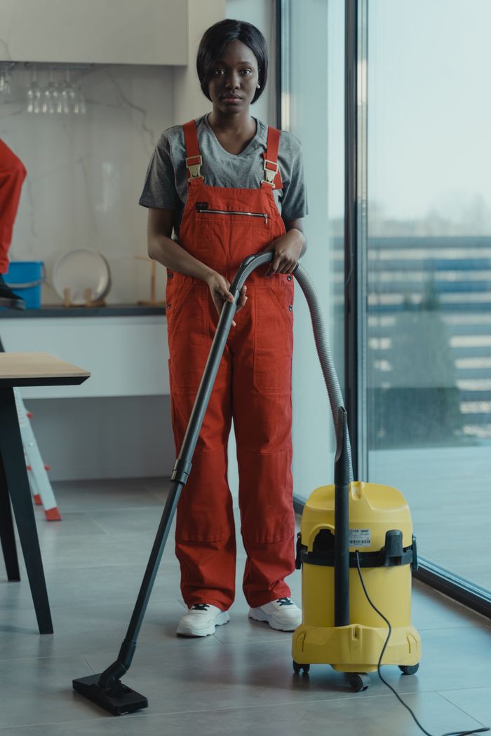 Une femme adulte en salopette rouge utilise un aspirateur dans un intérieur moderne, concentrée sur les tâches de nettoyage.